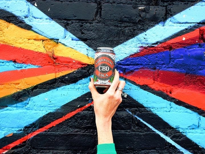 CBD Cola can held beside colorful painted brick wall