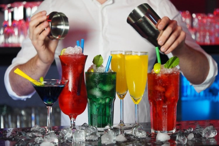 Six assorted cocktails on wooden bar beside bartender in white uniform