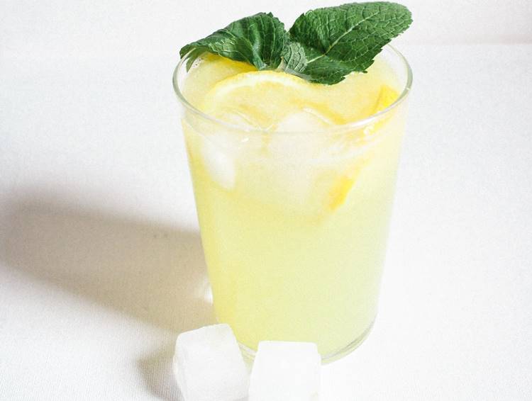 Tall iced lemonade glass mint leaf garnish beside ice cubes on white countertop