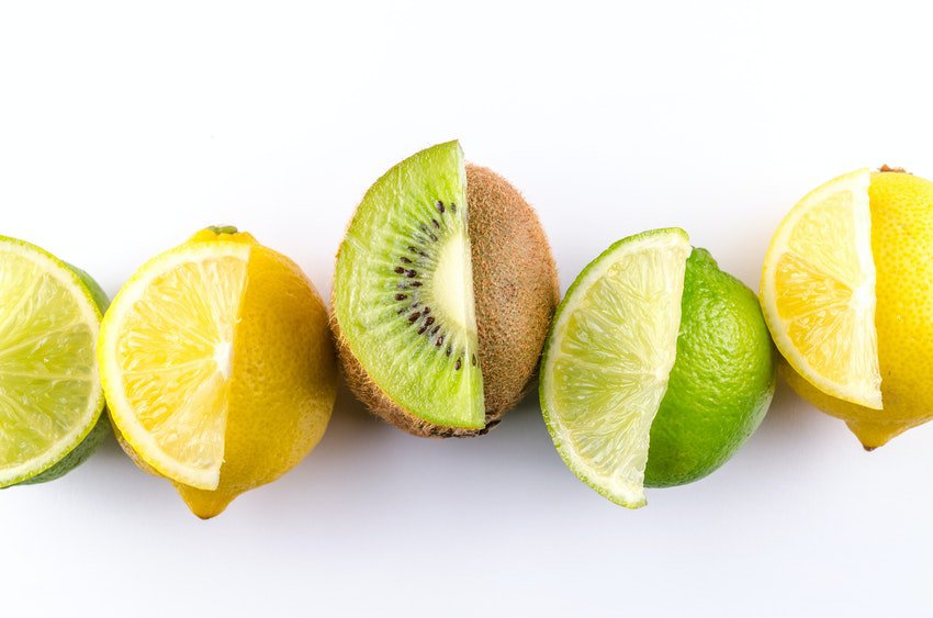 Whole limes, lemons and kiwis lined on flat, while surface, with one citrus slice on top of each fruit of the same type