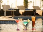 Three assorted cocktails in glasses on ground beside outdoor pool at dusk