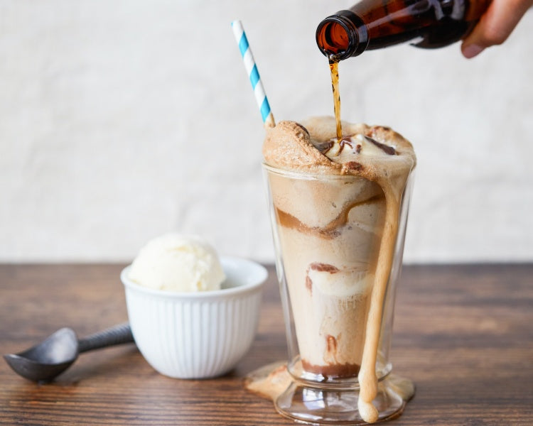 Bottled root beer pouring into iced cream dish with striped straw beside adjacent iced cream scooper