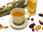 Mug of tea beside glass pitcher of iced tea, tea plants, hibiscus petals and herbs